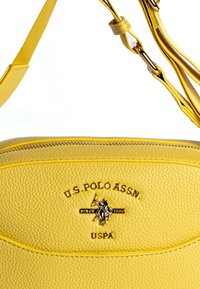 Borsa a tracolla gialla in pelle con superficie texture, con logo e dettagli in rilievo neri, e tracolla regolabile con hardware color oro.