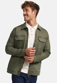 Grüne, mit Reißverschluss versehene Jacke mit zwei Brusttaschen, Hemdkragen und langen Ärmeln. Getragen über einem weißen Poloshirt und blauen Jeans, mit einer glatten Textur.
