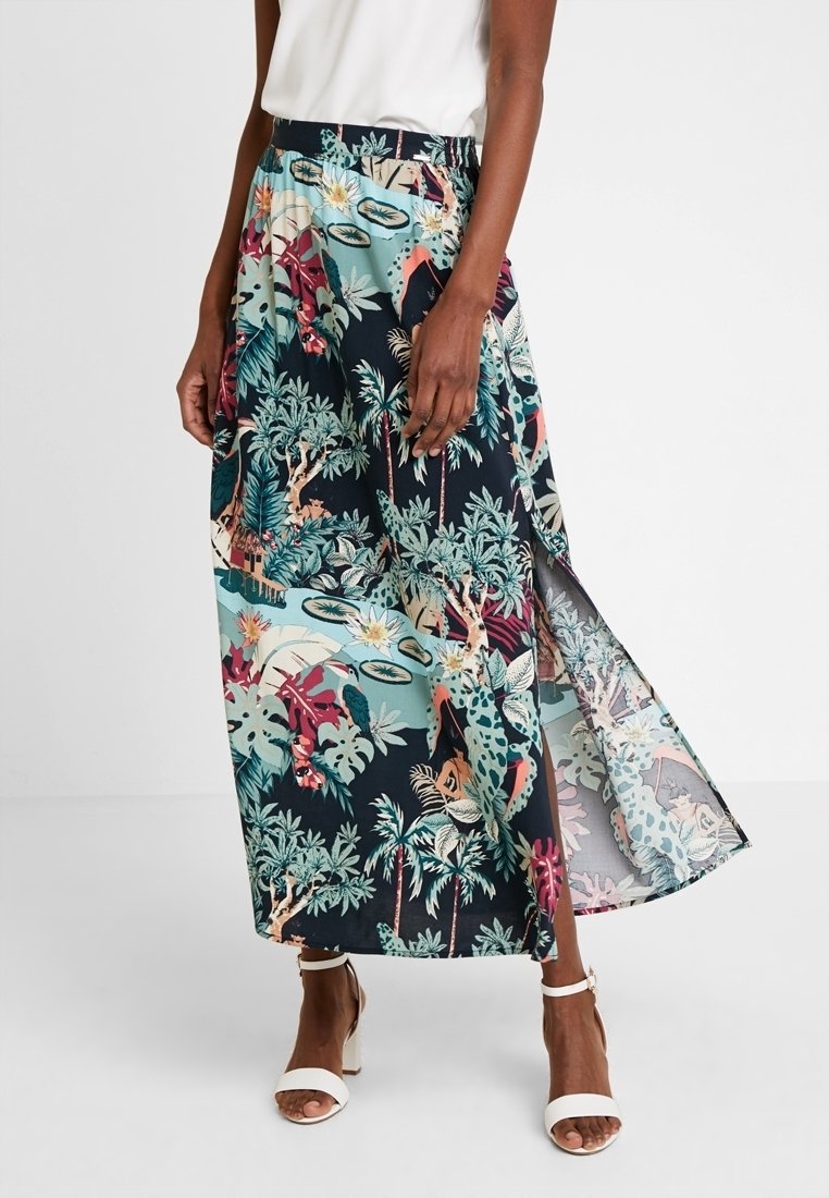 Femme portant une jupe longue à imprimé tropical coloré avec une fente latérale, un top blanc sans manches et des talons ouverts blancs à bride cheville.