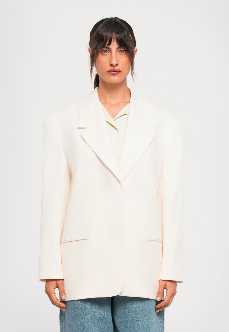 Vrouw met donker haar en een pony, gekleed in een oversized off-white blazer over een licht shirt en losse blauwe jeans tegen een effen witte achtergrond.