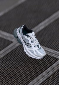 Scarpa atletica bianca e teal con tomaia in rete, colletto imbottito e suola in gomma spessa. Presenta un sistema di allacciatura e un logo distintivo sul lato.