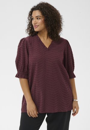 Woman with curly hair wearing a textured dark red blouse with puffed sleeves and black pants, looking to the side against a plain background.