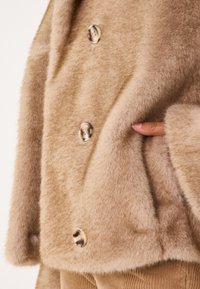 Veste en fausse fourrure couleur fauve avec de grands boutons clairs et des poches latérales. Texture douce et coupe décontractée, avec un large col.