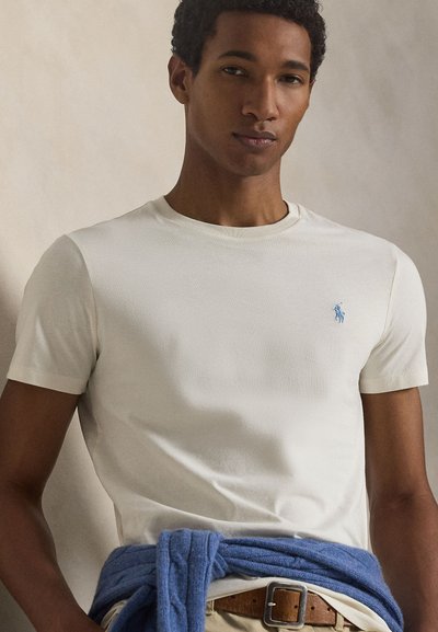 Mladý muž nosí jednoduché biele tričko s malým modrým logom, modrý sveter zviazaný okolo pása a béžové nohavice s hnedým opaskom.