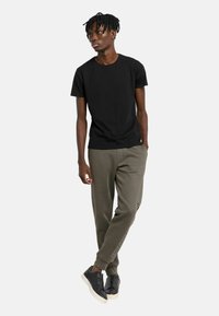 Schwarzes T-Shirt, olivgrüne Jogginghose mit seitlicher Beschriftung und schwarze Sneaker mit Reißverschluss-Detail. Einfaches, figurbetontes Design mit lässigem Look.