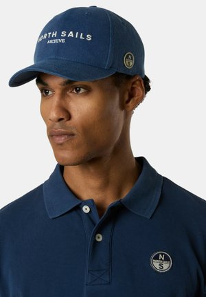 Uomo che indossa un cappellino North Sails blu navy e una polo abbinata con patch con il logo North Sails sul petto, guardando di lato.