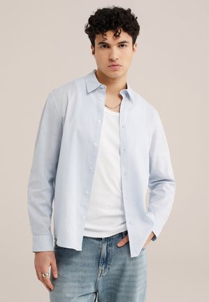 Młody mężczyzna o kręconych włosach, ubrany w jasnoniebieską koszulę na guziki na białej koszulce i jasnoniebieskich dżinsach, stoi na tle beżowego tła.