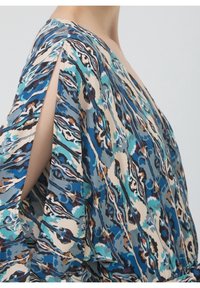 Femme portant un chemisier ample aux motifs multicolores bleu et beige, avec une fente verticale sur la manche dévoilant le haut du bras, sur un fond uni.