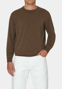 Maglione marrone lavorato a maglia con maniche lunghe e collo rotondo, caratterizzato da una texture liscia e polsini a coste, abbinato a pantaloni bianchi.