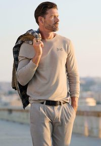 Beige Langarmhemd mit Logo, kombiniert mit passenden Hosen. Die Oberbekleidung hat ein strukturiertes grau-blaues Muster und liegt lässig über der Schulter.