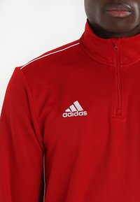 Rød atletisk quarter-zip topp laget av glatt stoff. Har hvite aksentstriper og et brodert hvitt Adidas-logo på brystet.