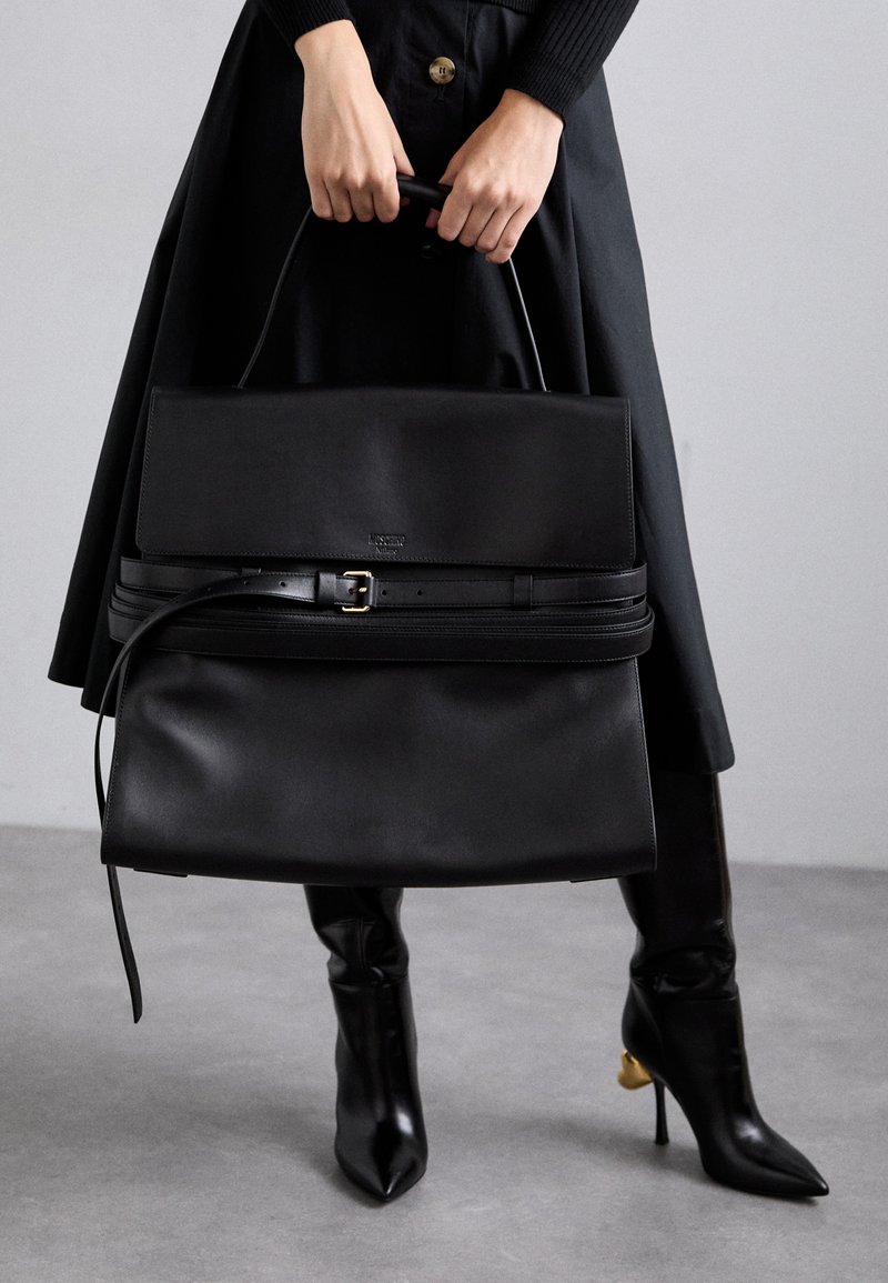 Persona in gonna nera e stivali, che tiene in mano una grande borsa in pelle nera con dettagli in cintura e manico superiore, su uno sfondo semplice.