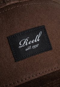 Parche de tela marrón con borde negro. Presenta texto bordado en blanco "Reell" y "est. 1997." Las texturas son suaves y mate.