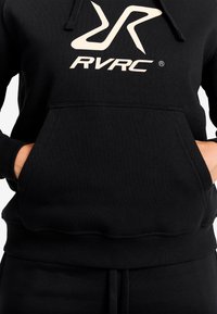 Svart hoodie med en framficka, försedd med en stor vit "RVRC"-logotyp. Materialet verkar mjukt med en texturerad yta och ribbade muddar.