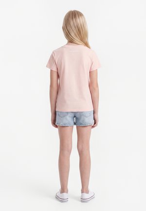 Dziecko o długich blond włosach, ubrane w jasnoróżowy t-shirt, jasne szorty jeansowe i białe trampki, stojące tyłem do kamery.