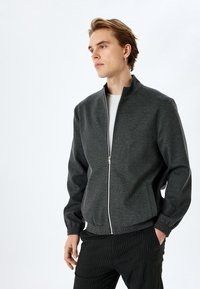 Grå zip-up jakke med høj krave og elastiske manchetter, der har et strømlinet design, for- og baglomme samt en glat tekstur, båret over en hvid skjorte.
