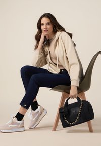 Felpa beige con zip e dettagli floreali, top beige, jeans scuri, sneakers sportive bianche e beige, e una borsa nera con dettagli in catena dorata.