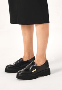Zwarte lakleren loafers met een chunky zool, voorzien van een decoratieve band en een gouden accent. Soepele textuur met een ronde teen ontwerp.
