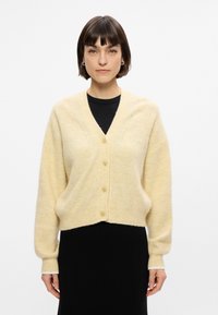 Cardigan à boutons en fil crème doux et texturé avec un col en V, doté de poignets et d'un ourlet côtelés, porté sur un haut noir.