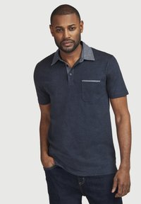 Marineblaues Polohemd mit einem kontrastierenden hellblauen Kragen, kurzen Ärmeln und Taschendetail. Hergestellt aus einem strukturierten Stoff. Getragen mit dunklen Jeans.