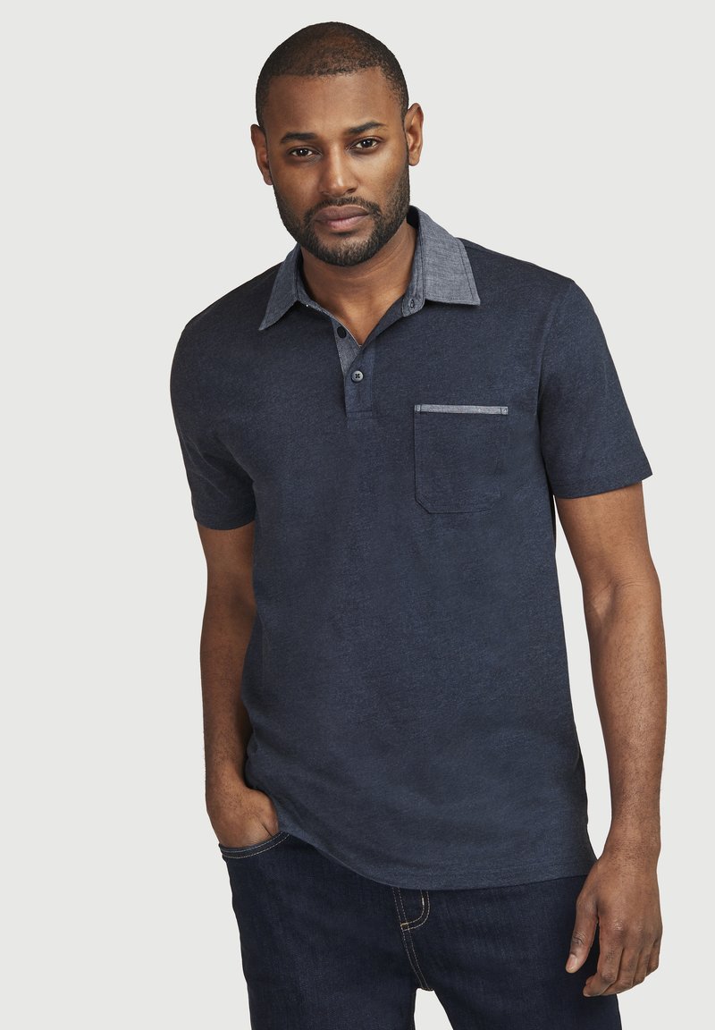 Marineblaues Polohemd mit einem kontrastierenden hellblauen Kragen, kurzen Ärmeln und Taschendetail. Hergestellt aus einem strukturierten Stoff. Getragen mit dunklen Jeans.