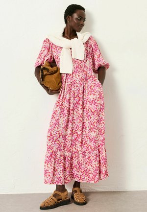 Frau in einem rosa, geblümten Maxikleid, weißem Pullover über den Schultern, hält eine braune Tasche und steht vor einer weißen Wand.