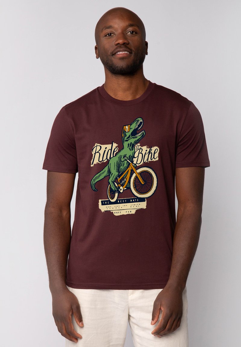 T-shirt bordeaux en coton, avec un graphique de dinosaure roulant à vélo et des accents textuels. Manches courtes et design classique à col rond.