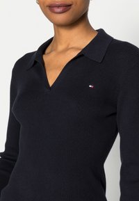 Marineblaues geripptes Polohemd mit V-Ausschnitt und langen Ärmeln. Mit einem kleinen gestickten Logo in Rot, Weiß und Blau auf der Brust.