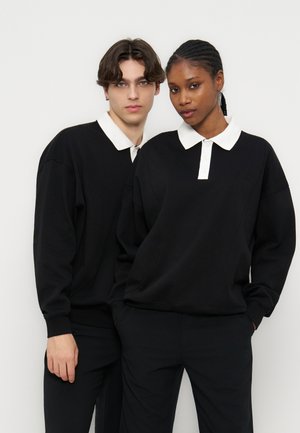 Twee personen dragen zwarte lange mouwen met witte kragen en staan dicht bij elkaar tegen een witte achtergrond, terwijl ze recht vooruit kijken.