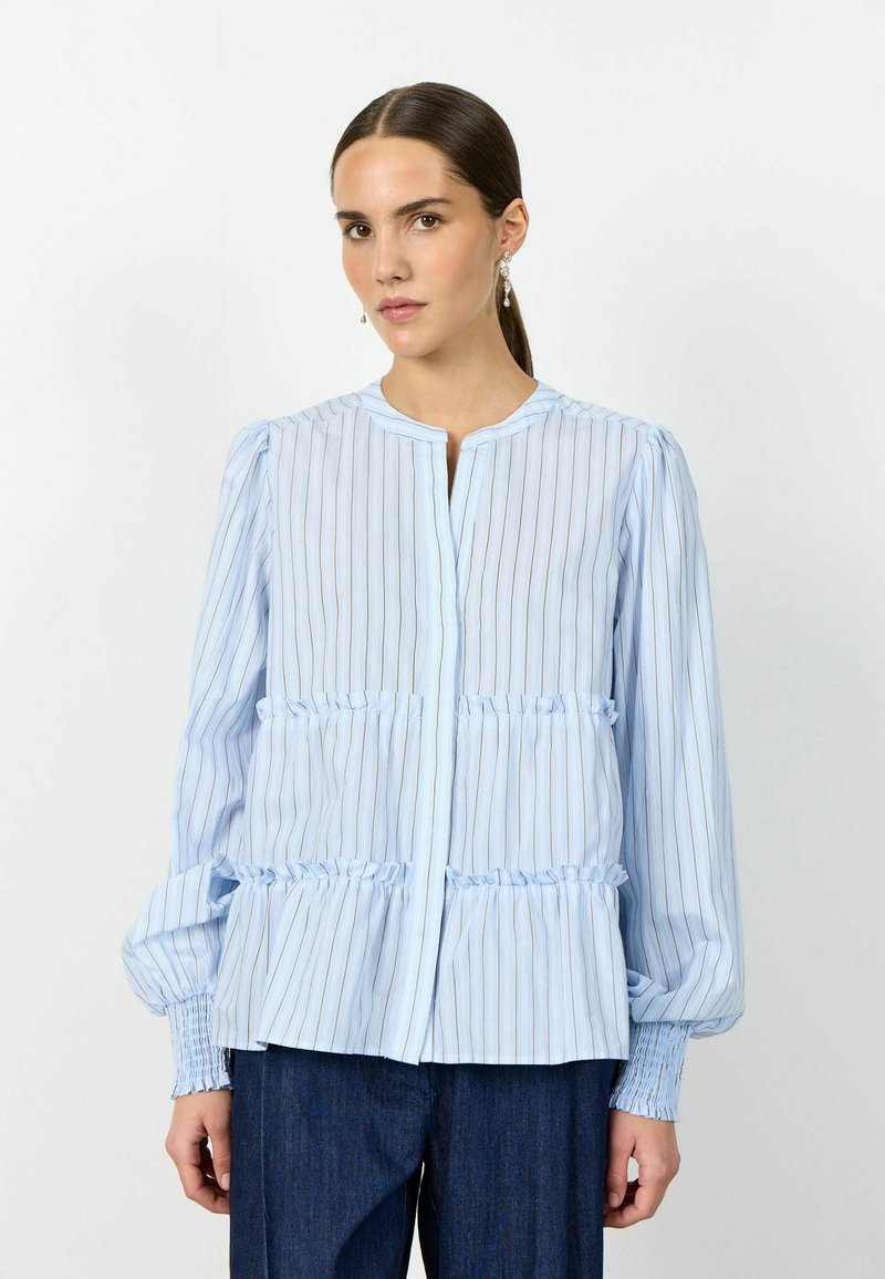 Vrouw draagt een lichtblauwe blouse met lange mouwen, gestreept en met geplooide lagen, en donkerblauwe broek, staat tegen een effen witte achtergrond.