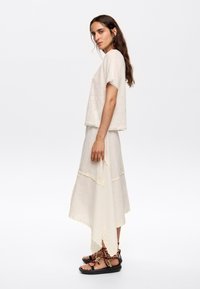 Cream short-sleeve top and layered maxi skirt with scalloped edges. Details include yellow stitching and brown strappy sandals.