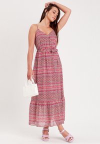 Robe maxi rose avec un motif géométrique, présentant des bretelles fines, une taille cintrée et une jupe à volants. Associée à des sandales lavande et un sac à main blanc.