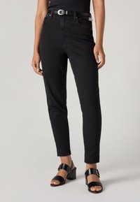Femme portant un jean skinny noir taille haute avec une ceinture noire et des sandales noires à talons, debout sur un fond clair.