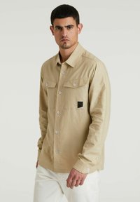 Beige Hemd mit langen Ärmeln, zwei Brusttaschen und einem schwarzen Logo-Patch. Der Stoff wirkt glatt mit einer leichten Textur.