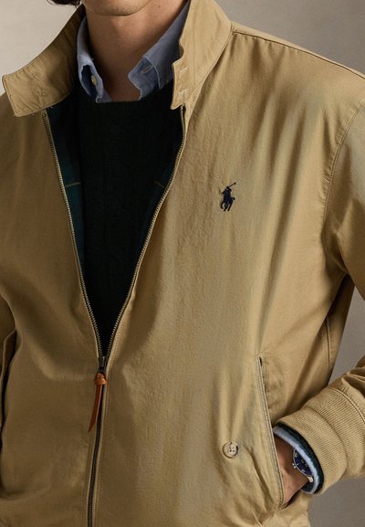 Mees kannab beežikat tõmblukuga jopet sinise kraega särgi ja musta kampsuni peal, käsi taskus ja nähtav käekell.