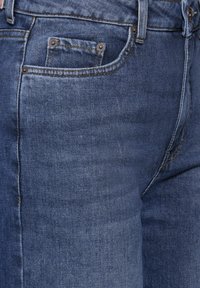 Vaqueros de mezclilla en azul oscuro con una ligera textura desgastada, que cuentan con cinco bolsillos, botones metálicos y costuras en contraste a lo largo de las costuras.