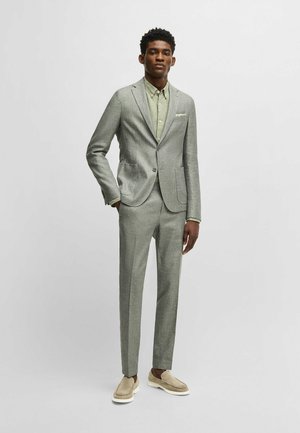 Un costume gris clair en tissu texturé avec une veste à deux boutons, des revers en notch et un pantalon droit. Assorti avec une chemise verte à col boutonné.