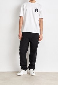 Weißes T-Shirt mit kurzen Ärmeln und einem schwarzen Blumenmotiv auf der linken Brust, kombiniert mit lockeren, schwarzen Cargo-Hosen und weißen Sneakers.