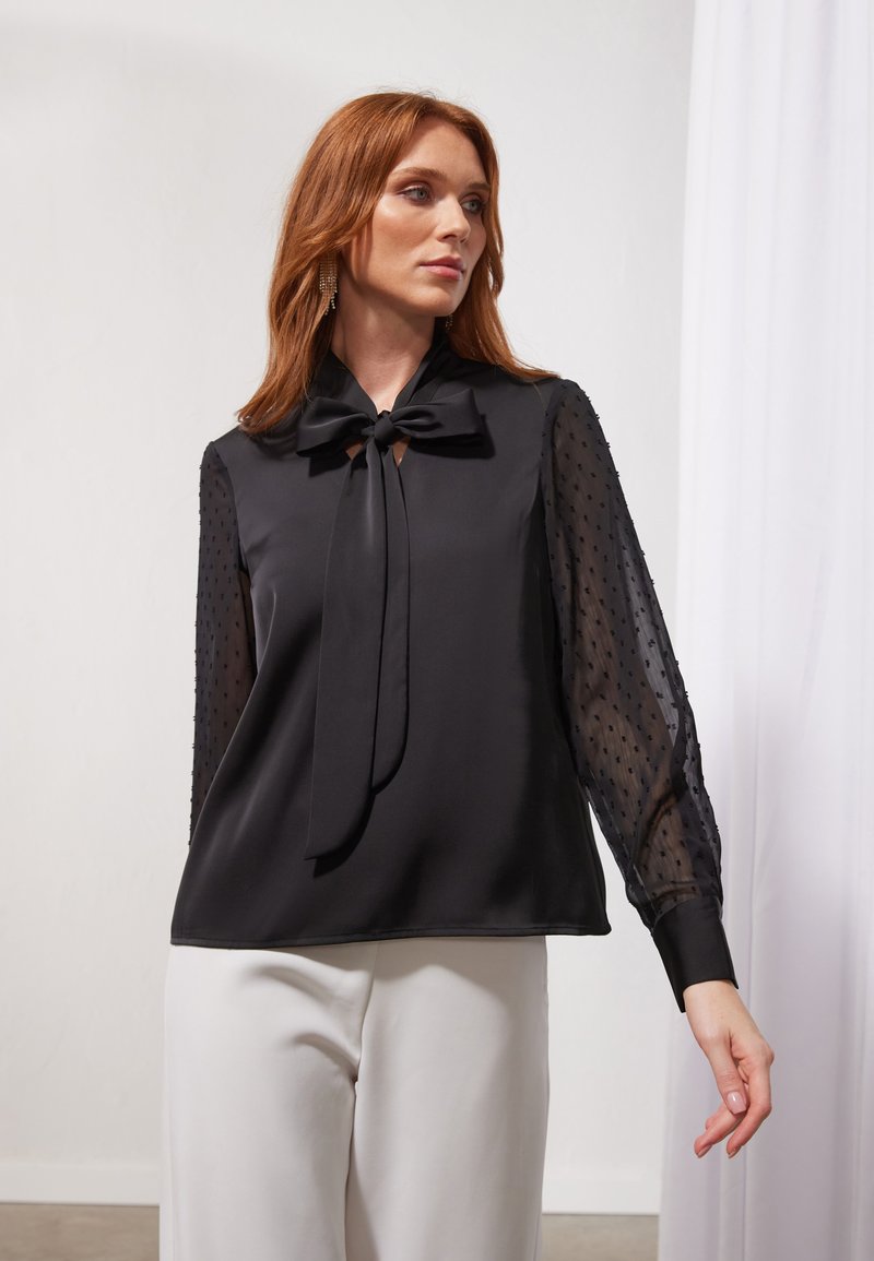 Black blouse with a necktie, featuring solid satin front and sheer, dotted long sleeves. Smooth texture and relaxed fit.