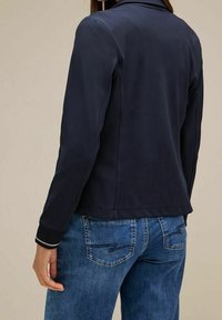 Námořnická modrá bunda s hladkou texturou, s límečkem a kontrastními manžetami; kombinována se světle modrými džíny s designem zadní kapsy.