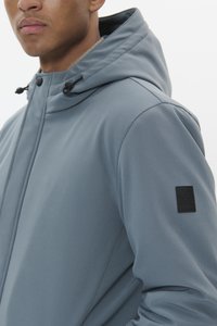 Siva kapuca jakna iz gladke tkanine, ki prikazuje posnetek ovratnika in dekorativno nalepko na rokavu.