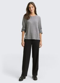 Woman wearing a patterned grey long-sleeve top, black wide-leg trousers, and black platform shoes, standing against a plain background.