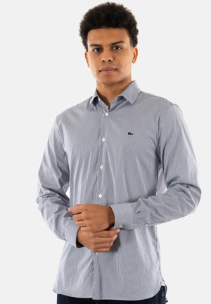 Camisa de botones a rayas en azul y blanco, tela texturada, mangas largas, corte entallado, con un pequeño logo en el pecho.