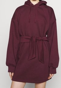 Weinrotes Kapuzenkleid mit langen Ärmeln, einem gebundenen Taillengürtel und gerippten Bündchen. Glatter Stoff mit lässiger Passform.