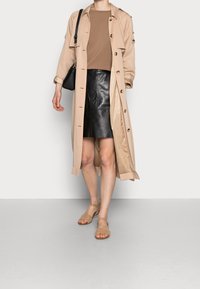 Eine Frau trägt einen beigen Trenchcoat, ein braunes Oberteil, einen schwarzen Lederrock, beige Sandalen und hält eine schwarze Schultertasche. Sie steht vor einem schlichten Hintergrund.
