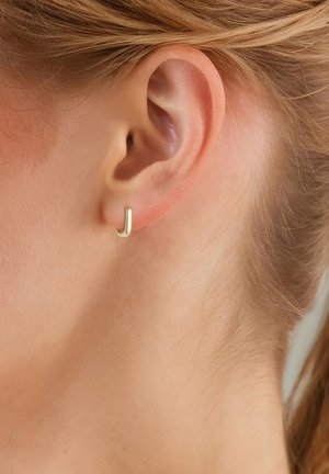 Gouden oorring met een glad, minimalistisch ontwerp, weergegeven op een close-up van een oor tegen lichte huid en steil haar.