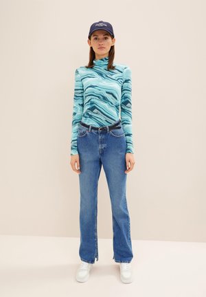 Langärmliges Oberteil mit einem blauen und türkisfarbenen wellenförmigen Marmor-Muster, kombiniert mit geraden blauen Jeans, ergänzt durch eine marineblaue Kappe und weiße Sneakers.