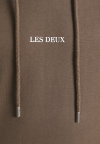 Tejido de sudadera marrón con el texto blanco "LES DEUX" centrado, presentando cordones a juego con extremos metálicos de color plateado.