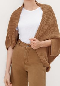 Bruine gebreide sjaal gedrapeerd over een wit T-shirt, gecombineerd met hooggetailleerde bruine stoffen broek. Voorzien van een geribbelde textuur en subtiele stiksels.