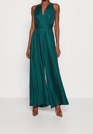 Bezrękawowy turkusowy satynowy kombinezon z szerokimi nogawkami, głębokim dekoltem w kształcie litery V oraz cienkim wcięciem w talii, noszony z cielistymi szpilkami z wąskim noskiem.